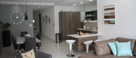 A modern living room with a brown leather sofa, a dining area with a glass table, and a kitchen with a white countertop.