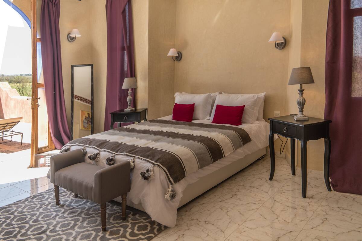 A bedroom with a bed, two bedside tables, a chair, and a view of the outdoors.