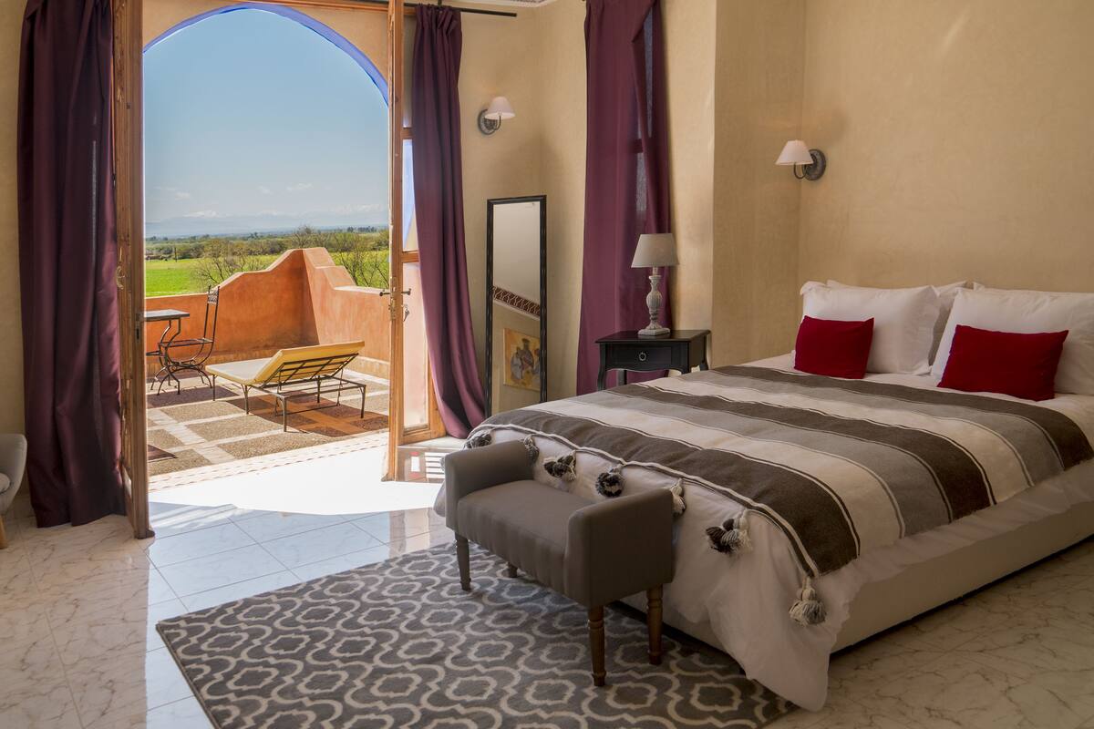 A bedroom with a large bed, a bench, a nightstand, a lamp, and a view of the outdoors.