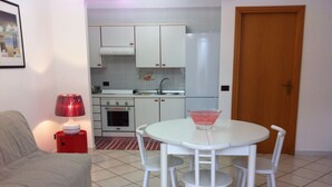 Une cuisine de taille réduite, avec des meubles blancs, une cuisinière et un évier. On y trouve une table à manger avec quatre chaises, ainsi qu’une petite table d’appoint rouge sur laquelle est posée une lampe. Une porte en bois est visible au fond.