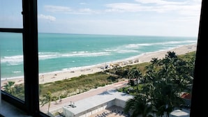 Vista di una spiaggia da una finestra, con onde, palme e un edificio a tetto piatto.