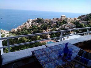 A balcony with a view of a coastal town, a table with a patterned tablecloth, and blue glassware.