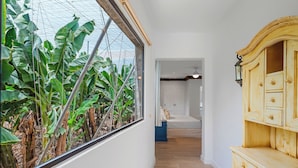 Un pasillo con vistas a plantas de banano a través de una ventana, un armario de madera y una cama al fondo.