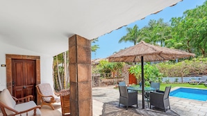 Un patio con piscina, sillas de madera, una mesa y un paraguas de paja.
