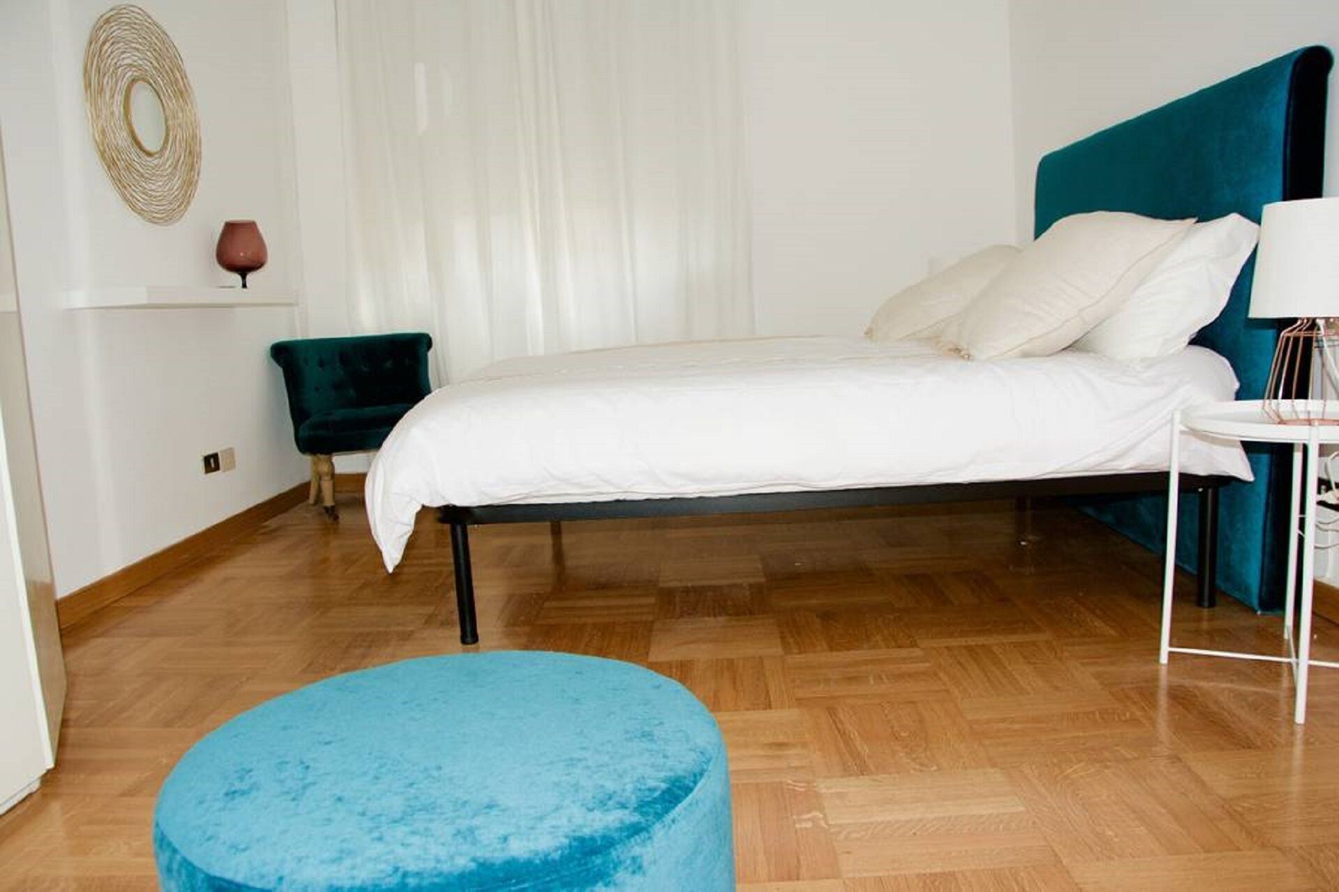 Ein Schlafzimmer mit einem Bett, einem blauen Hocker, einem Nachttisch, einer Lampe und einer Wanddekoration.