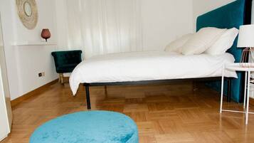 Ein Schlafzimmer mit einem Bett, einem blauen Hocker, einem Nachttisch, einer Lampe und einer Wanddekoration.