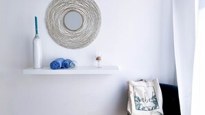 A room with a white wall, a round woven mirror, a white shelf with a blue bottle, rolled towels, and a glass. There's a teal sofa and a white bag with text. - Flaminio Penthouse with Spectacular Terrace (Roma)