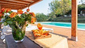 A poolside setting with a wooden pergola, a glass vase with orange roses, a glass of orange juice, and a basket of snacks.