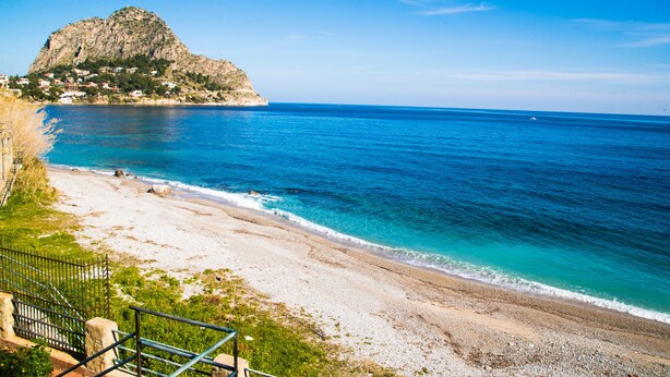 A coastal view with a rocky mountain, clear blue sea, and a sandy beach.