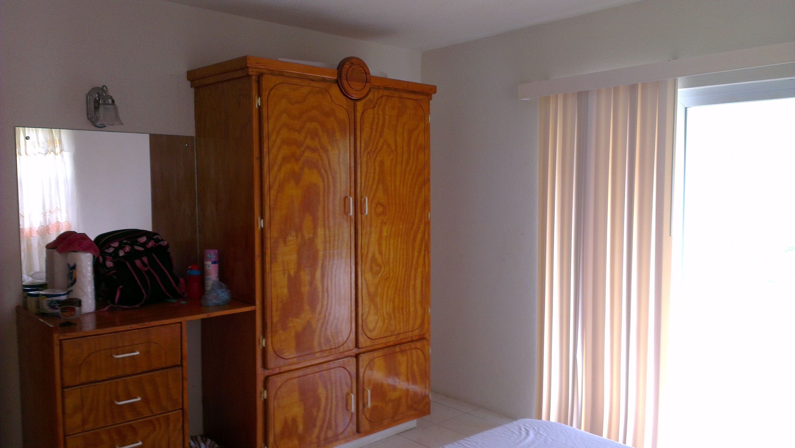 Een slaapkamer met een houten garderobe, een bed, een raam met gordijnen en een spiegel aan de muur.