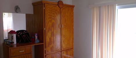 Een slaapkamer met een houten garderobe, een bed, een raam met gordijnen en een spiegel aan de muur.