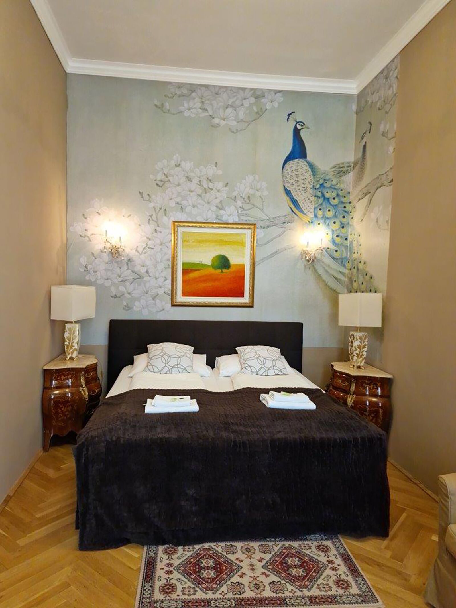Ein Schlafzimmer mit einem großen Bett, zwei Nachttischen mit Lampen, einem Wandbild von einem Pfau und einem gerahmten Gemälde über dem Bett.