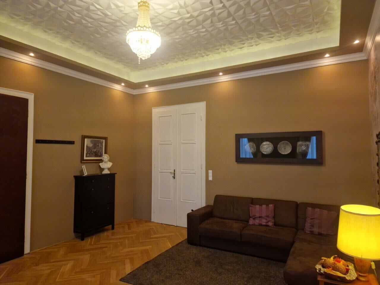 A living room with a herringbone floor, a brown sofa, a black cabinet, a white door, and a chandelier.