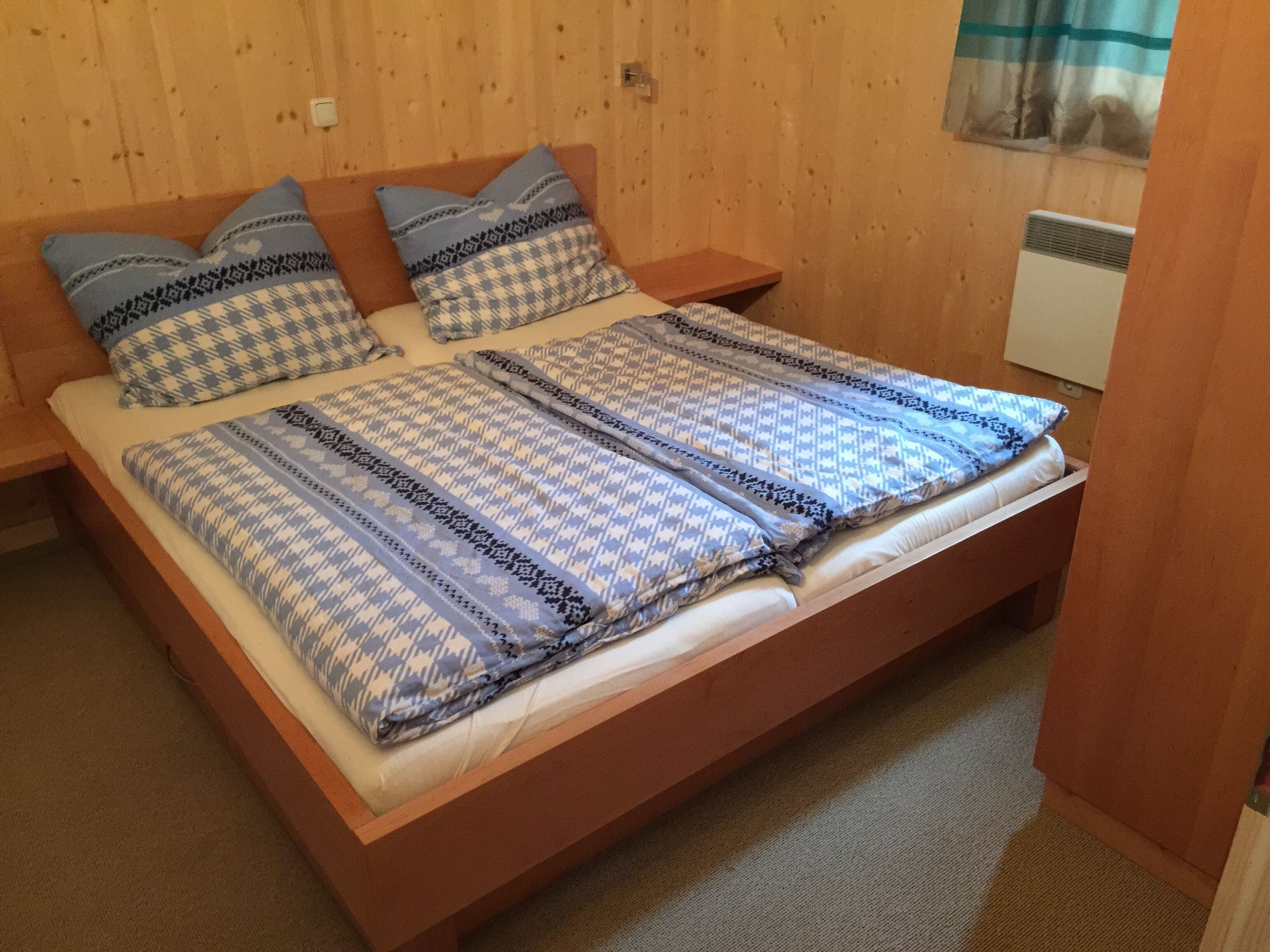 A wooden bed with a patterned bedspread and pillows, a nightstand, and a window with curtains.