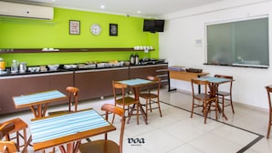 A hotel kitchen with a green wall, a clock, and a flat-screen TV. There are tables and chairs arranged for dining. - VOA Convenience Hotel (Jundiai)