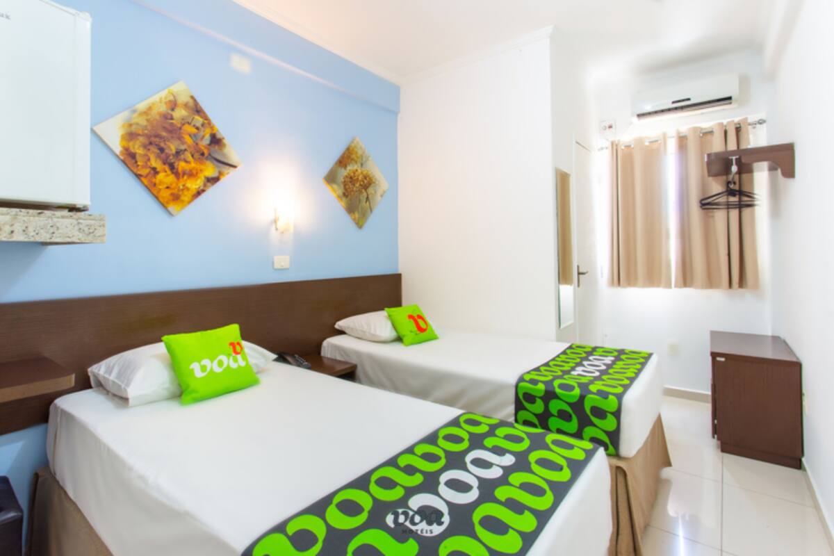 A hotel room with two beds, each with a green and black patterned bedspread, white pillows, and a small green pillow with a red 'B' on one bed. There are two wall-mounted paintings, a white air conditioning unit, a wooden cabinet, and a window with beige curtains.