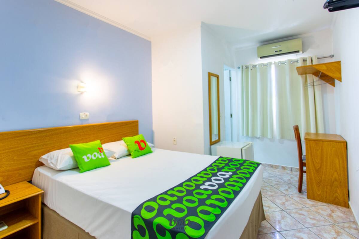 A hotel room with a bed, a nightstand, a wooden headboard, a green and black patterned bedspread, and a white wall with a light switch.