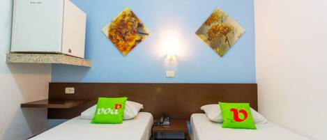 Quarto de hotel com duas camas, uma escrivaninha, uma prateleira com geladeira e duas obras de arte penduradas na parede.