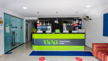 Un mostrador de recepción de hotel con una decoración en tonos verde brillante y gris, una pantalla digital y una zona de espera con un felpudo azul y un sofá a rayas.