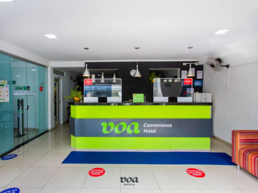 Un mostrador de recepción de hotel con una decoración en tonos verde brillante y gris, una pantalla digital y una zona de espera con un felpudo azul y un sofá a rayas.