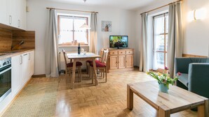Moderni keittiö, jossa on puiset kaapit, sekä ruokailutila, jossa on pöytä ja tuolit. Olohuoneessa on televisio jalustalla ja sohva.