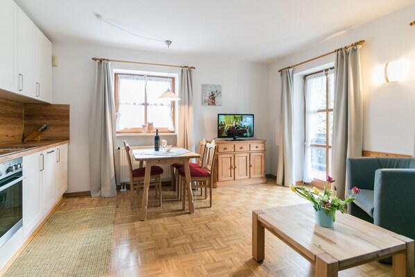 Eine moderne Küche mit Holzschränken und einem Essbereich mit Tisch und Stühlen. Ein Fernseher steht auf einem Schrank und im Wohnbereich befindet sich eine Couch.