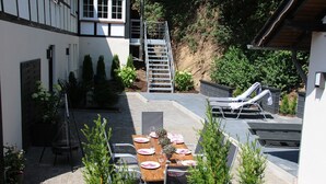 Un patio con una mesa de comedor preparada para cuatro personas, rodeada de sillas, una escalera de metal y una zona de estar con un sofá y una butaca.