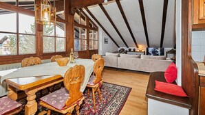 A dining area with a wooden table and chairs, a kitchen area with a sofa, and a view of the outside through large windows. - Dachgeschoss-Ferienwohnung (5) (Reit im Winkl)