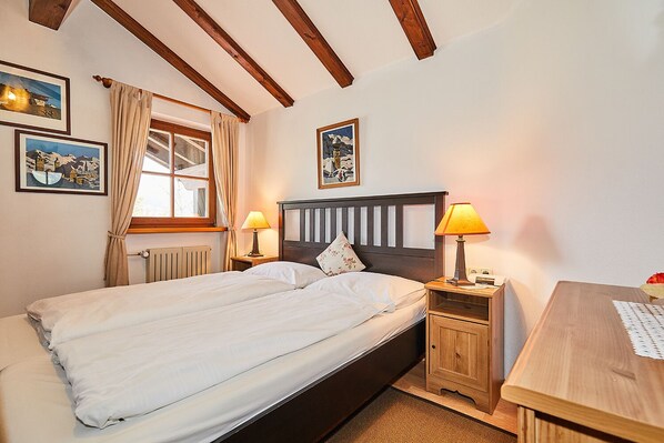 A bedroom with a double bed, two bedside tables with lamps, a wooden desk, and a window with curtains. - Dachgeschoss-Ferienwohnung (5) (Reit im Winkl)