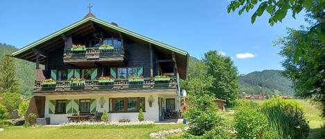 A two-story wooden chalet with balconies, a garden in front, and a mountainous backdrop.