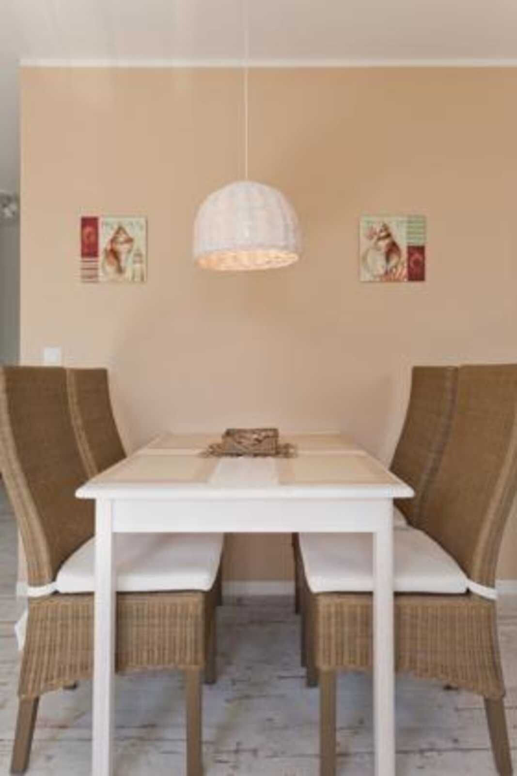 Un comedor con una mesa blanca, sillas de mimbre, una lámpara colgante y dos carteles en la pared.