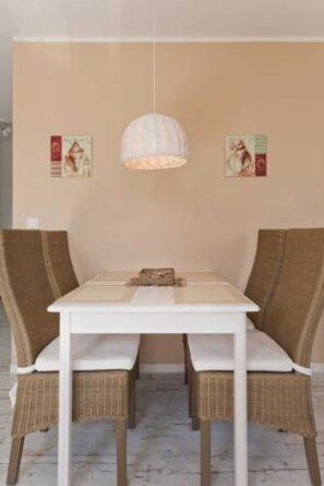 A dining area with a white table, wicker chairs, a hanging light fixture, and two wall-mounted posters. - FeWo Strandperle A 2. 04 Ref. 128693 u 1 - House Seaview Apartment Strandperle A 2. 04 Ref. 128693 u 1 (Baabe)