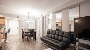 Un salon moderne avec un canapé en cuir, une table à manger et des chaises. On aperçoit un téléviseur et une grande fenêtre au fond.