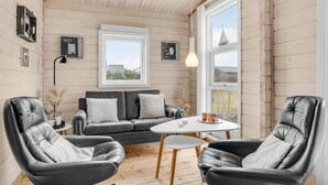 Et hyggeligt rum med en lædersofa, to læderlænestole, et lille rundt bord og et stort vindue med udsigt til en sejlbåd.