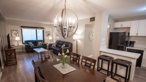 A modern living room with a dining table, sofa, and kitchen area with a refrigerator and coffee maker.