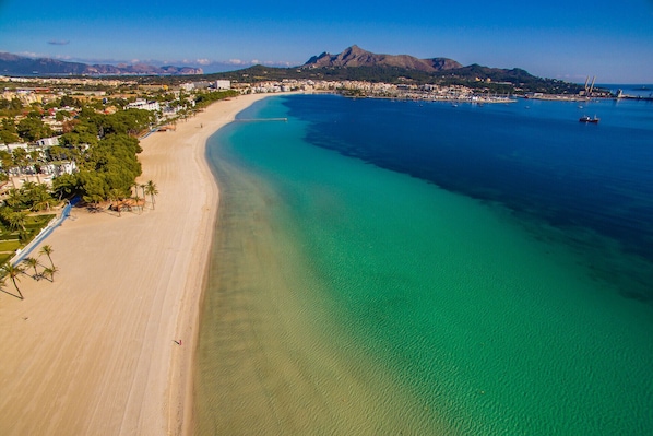 An aerial view of a coastal area with a sandy beach, clear blue waters, and a mountainous backdrop.
