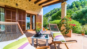 A patio with two wooden lounge chairs, a small table with drinks, and a view of a lush garden and hills.