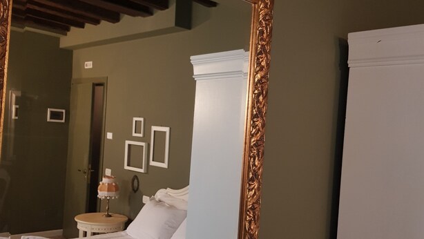 A hotel room with a large ornate mirror, a bed with white linens, a nightstand with a lamp, and a door in the background. - Casa Fornaretto (Venice)