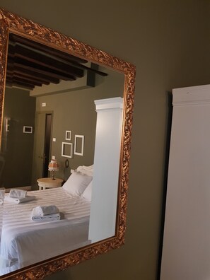 A hotel room with a large ornate mirror, a bed with white linens, a nightstand with a lamp, and a door in the background. - Casa Fornaretto (Venice)