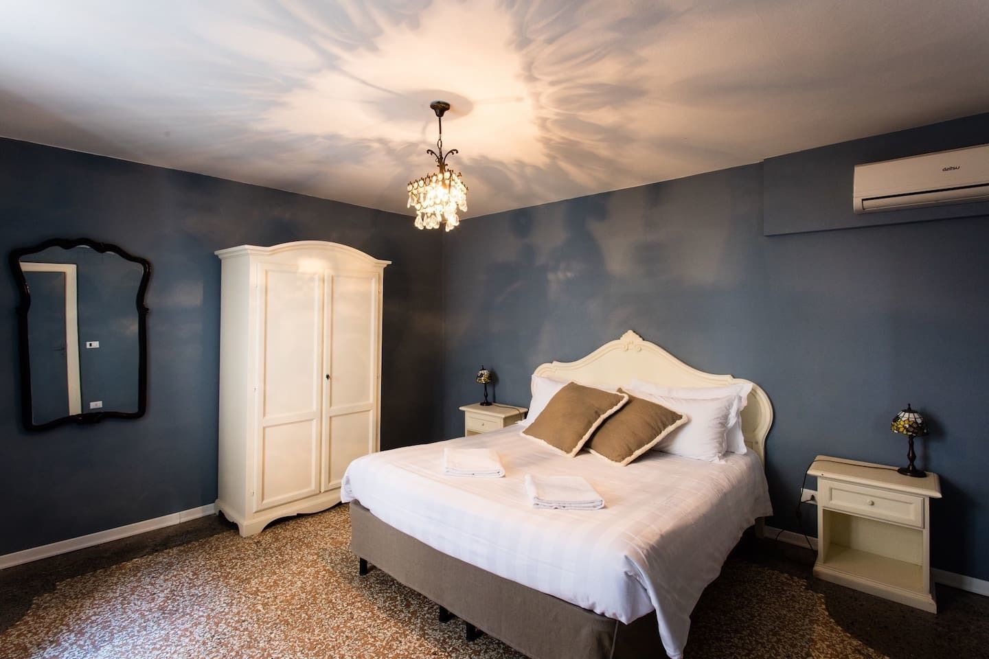 A bedroom with a bed, nightstands, a wardrobe, a chandelier, and an air conditioning unit.