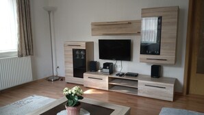 Een moderne woonkamer met een flatscreen televisie, een houten tv-meubel en een potplant op een bijzettafeltje.