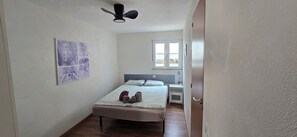 Een slaapkamer met een bed, een nachtkastje, een plafondventilator, een raam en een aan de muur hangend kunstwerk.