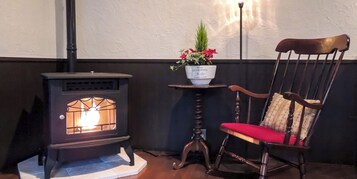 Una estufa de leña, una mecedora de madera con cojín rojo, y una mesita con una planta en maceta.
