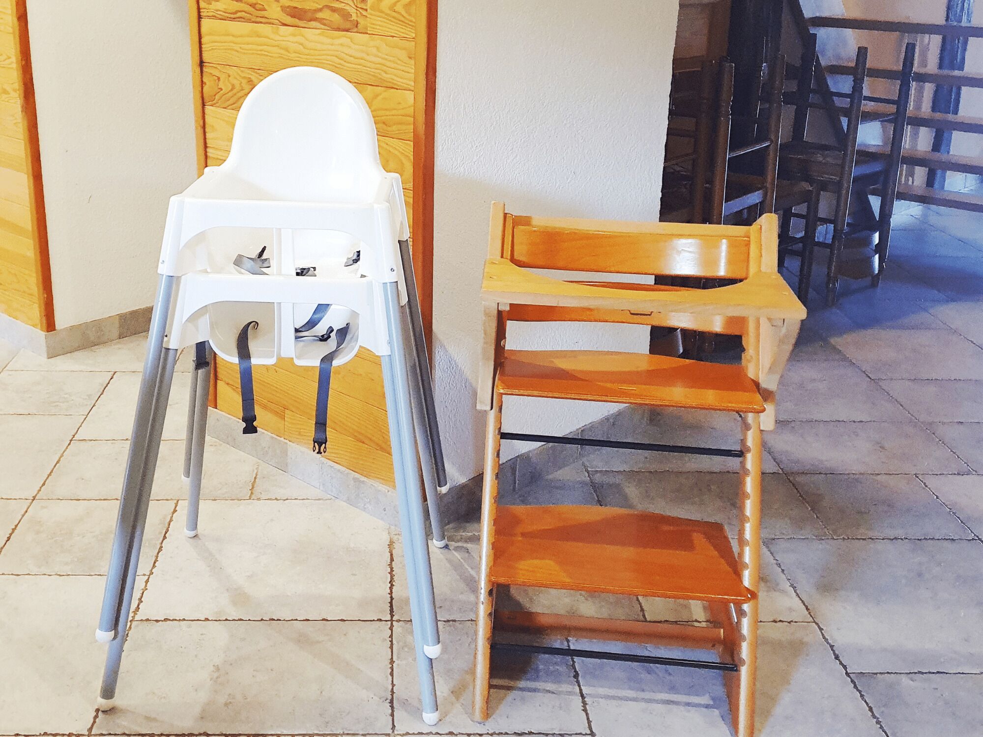 A high chair and a wooden chair in a room with tiled flooring and wooden paneling on the walls.