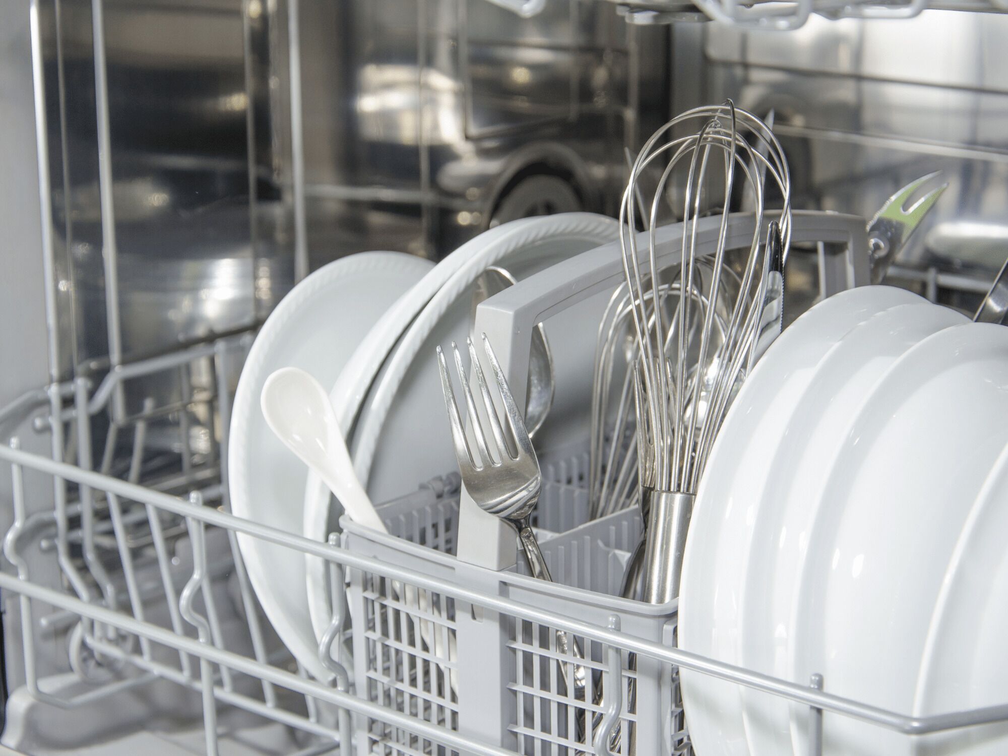 A dishwasher filled with white dishes, forks, and a whisk.