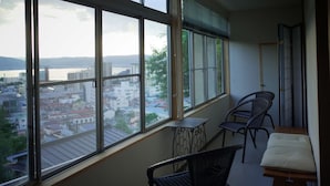A balcony with wicker chairs, a small table, and a view of a cityscape and water. - chambre d'hôte TATAMI - Hostel (Suwa)