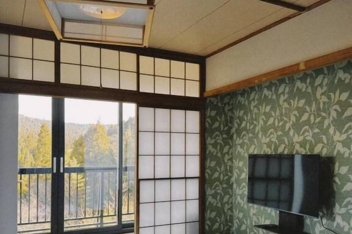 Ein Hotelzimmer mit Tatami-Boden, einer Schiebetür zum Balkon, einem an der Wand befestigten Fernseher und einem Bett mit weißer Bettwäsche.