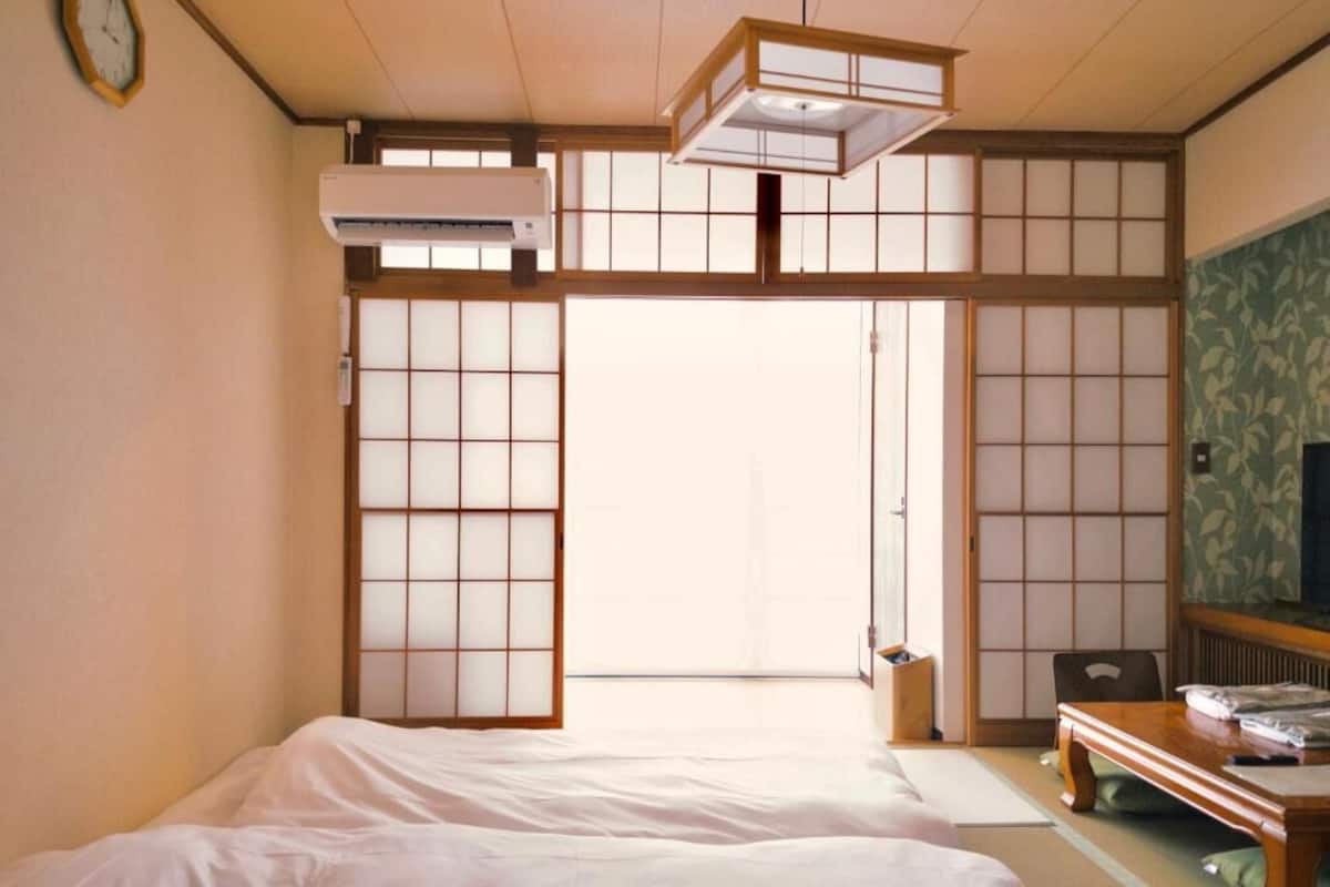 Ein traditioneller japanischer Raum mit Tatami-Böden, einem niedrigen Holzbett, einer gleitenden Shoji-Schiebetür und einem kleinen Schreibtisch mit Stuhl.