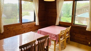 A wooden cabin interior with a dining table and chairs, large windows with curtains, and a hanging light fixture. - Yado Brodiaea (Myoko)
