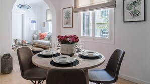 Un comedor con una mesa redonda dispuesta para cuatro personas, rodeada de sillas. Hay un florero con flores sobre la mesa, y una ventana con persianas a un lado.
